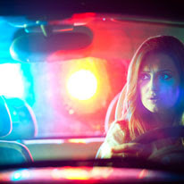 A woman looks in her car's rear-view mirror as police lights flash in the background. She is being pulled over.
