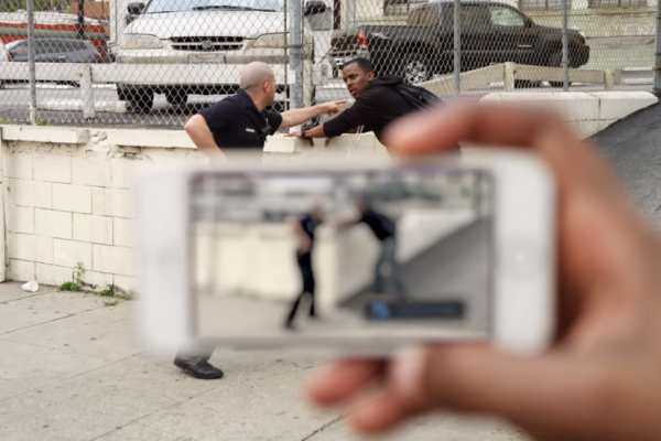 A police officer is gesturing at a man in the background. In the foreground, a hand is holding a phone recording the encounter.