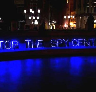 Blue lights in Oakland plaza at night spell out STOP THE SPY CENTER