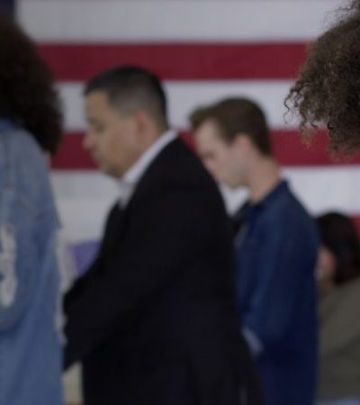 Young woman of color staffing desk at polling station with various voters in background, US flag on wall behind them.