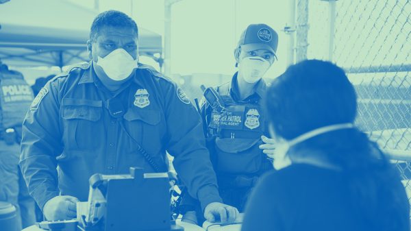 Customs and Border Patrol officer questioning a person at a port of entry. Both are wearing face masks.
