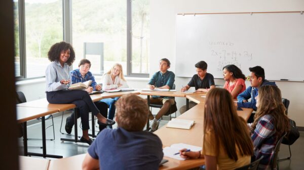 Female High School Tutor Sitting At Table With Pupils Teaching Class