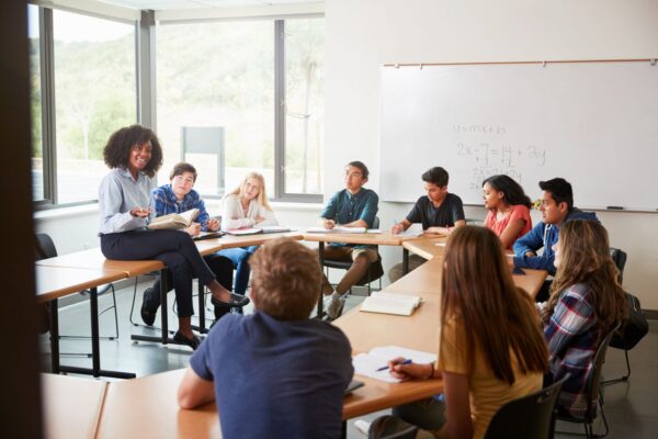 Female High School Tutor Sitting At Table With Pupils Teaching Class