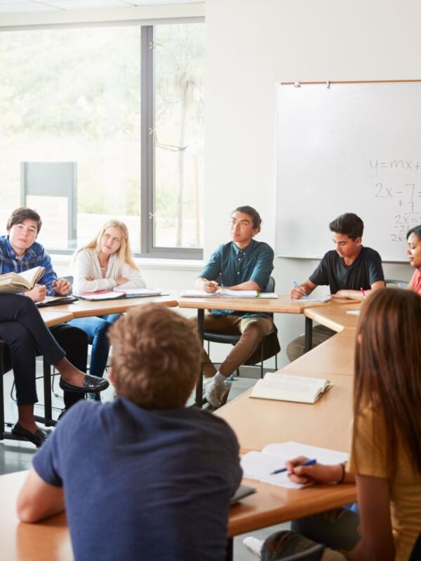 Female High School Tutor Sitting At Table With Pupils Teaching Class