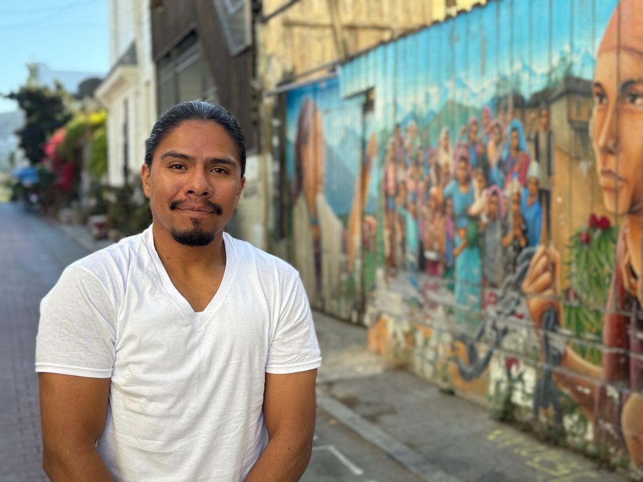 Picture of Jose Ruben. Young, latino man wearing a white t shirt. He is standing next to a colorful mural