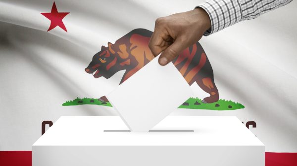 A hand placing a ballot into a ballot box with the California state flag in the background.