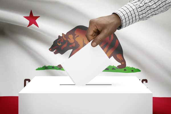 A hand placing a ballot into a ballot box with the California state flag in the background.