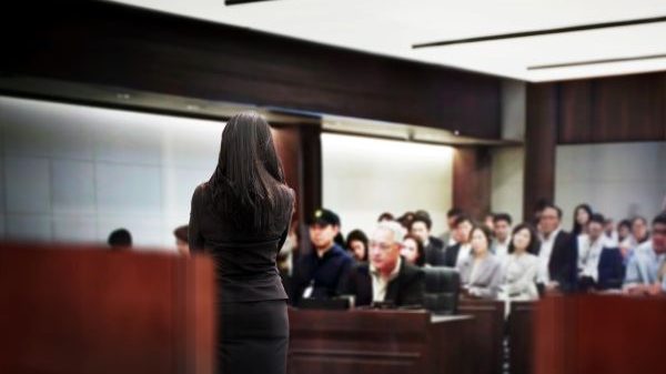 Court room with a person standing and several individuals seated in rows behind observing the proceedings.
