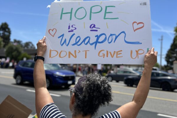Photograph of a person from the back holding a protest sign saying "Hope is a weapon. Don't give up."