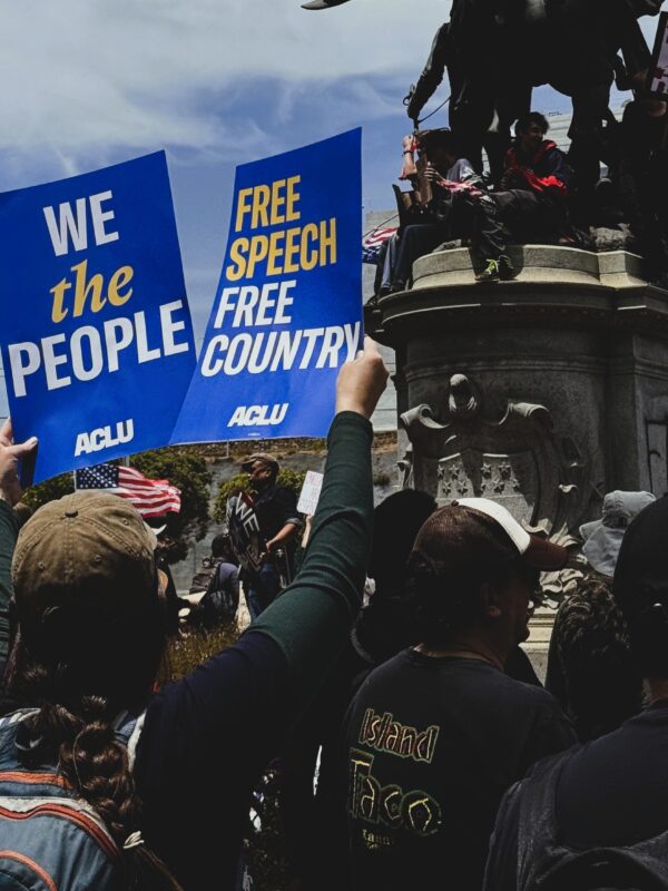 Photo of a people at a protest holding up ACLU branded signs saying, 'We the People' and 'Free Speech, Free Country.'
