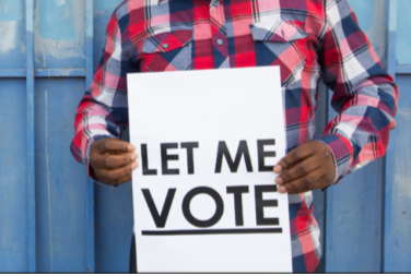torso and hands of a man in a checked shirt holding a sign that says Let Me Vote