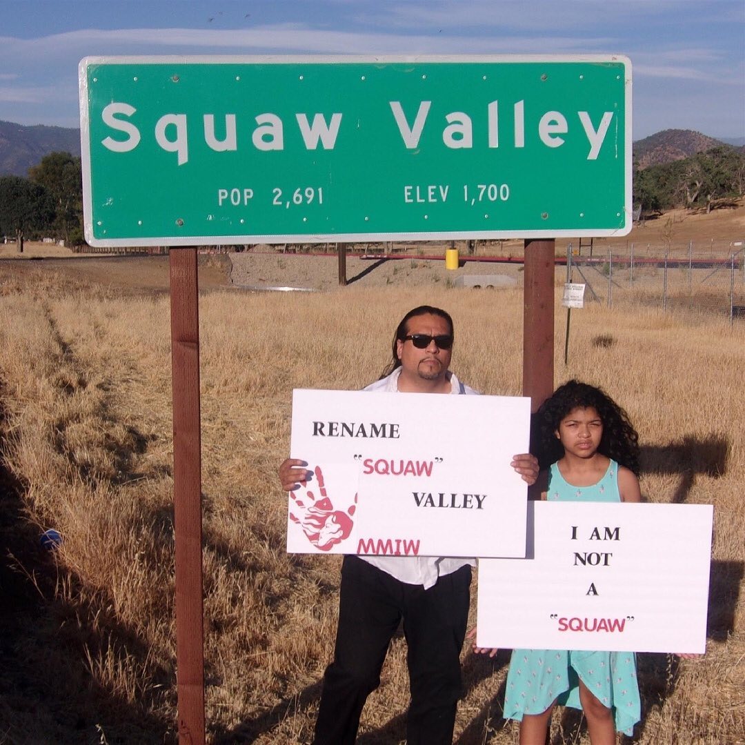Indigenous advocate Roman C. Raintree and his daughter stand in front of a road sign that reads "Squaw Valley". The two are holding posters - one that reads "Rename Squaw Valley" and the other says "I am not a Squaw"